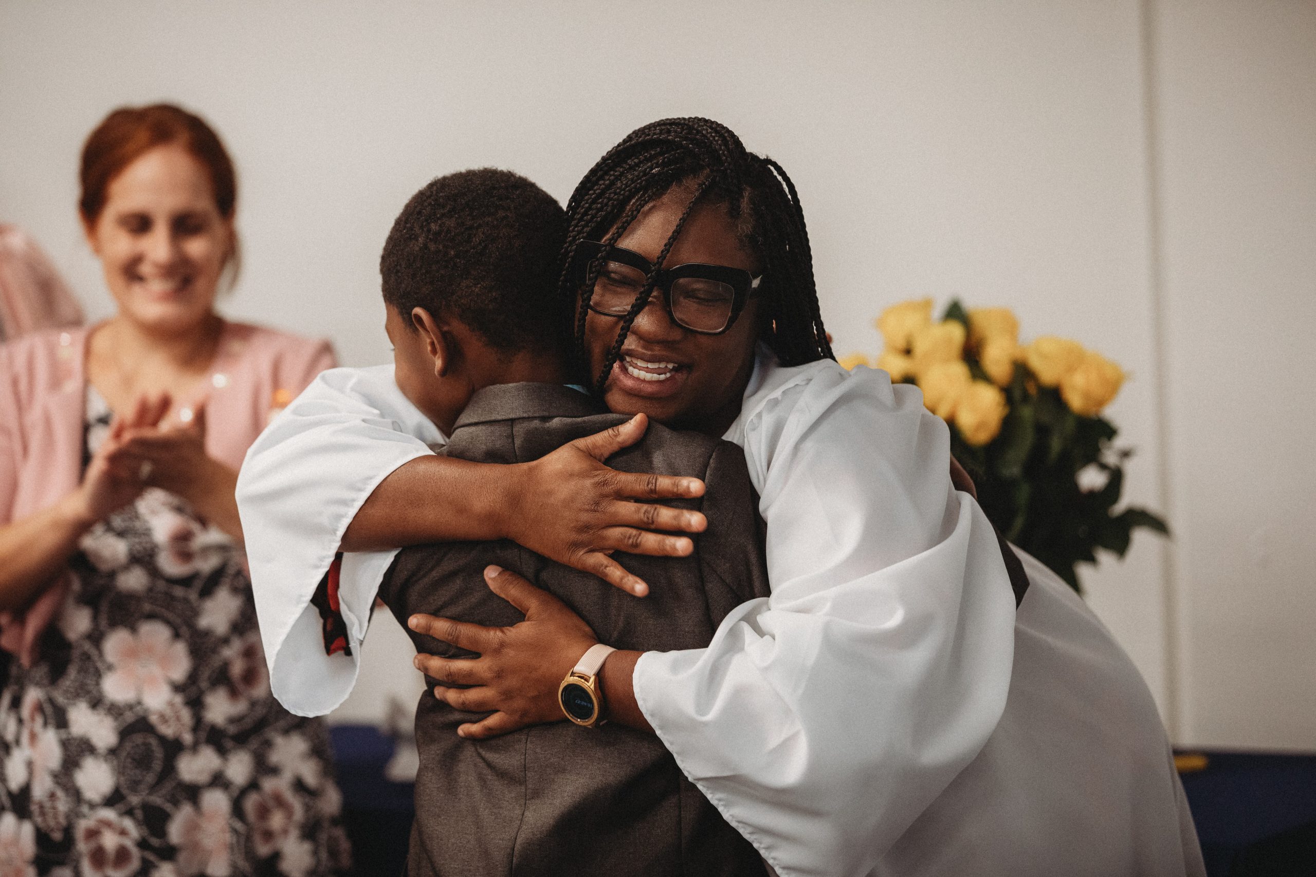 A smith chason college graduate is in her cap and gown smiling big and hugging her young son. Behind her is a Smith Chason staff member smiling and clapping in celebration of the new grad